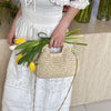 A woman holds a woven straw chain crossbody handbag with green onions and garlic stalks in a white dress.
