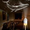 A white projector casts a glowing whale silhouette onto the ceiling in a dark bedroom.