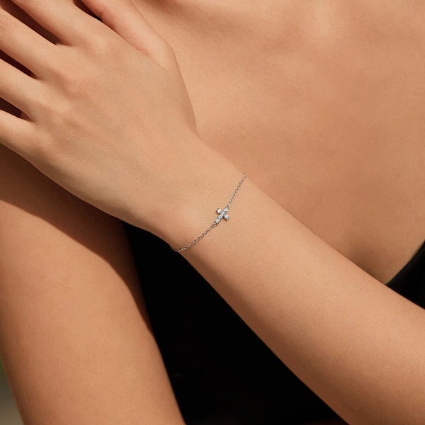 A close-up of a hand wearing a delicate silver bracelet featuring a minimalist double cross design.