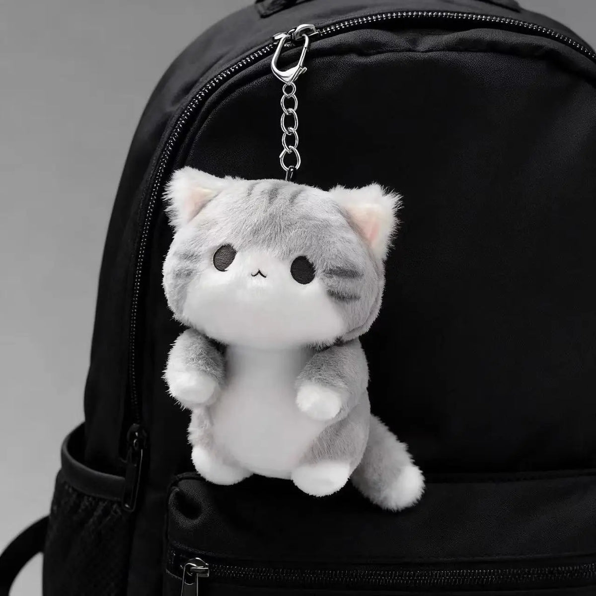 A soft grey cat plush charm attached to a black backpack zipper.