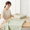 A woman is folding and storing a light green bedding item inside a clear mesh storage bag on a wooden table.