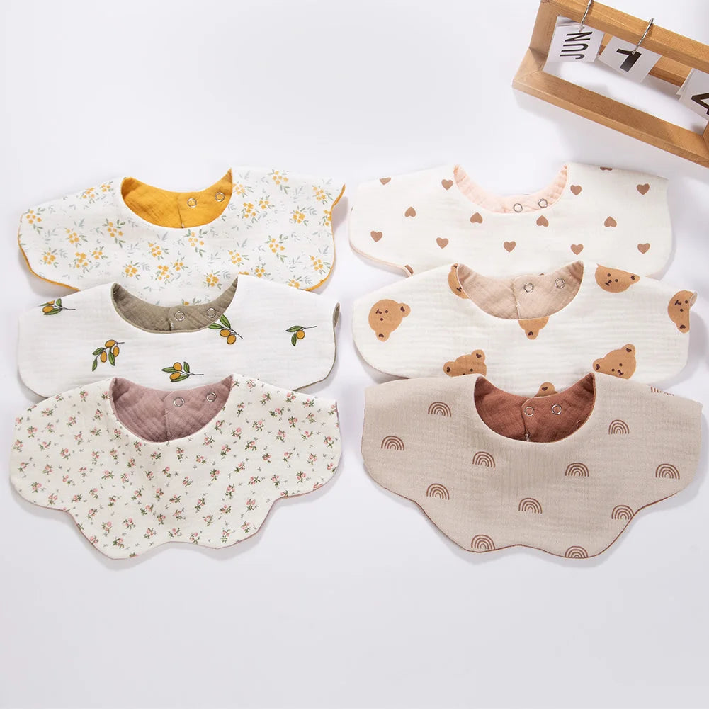 A collection of light-colored muslin cotton baby bibs and burp cloths with various patterns and snap closures.