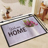 A rectangular minimalist geometric non-slip absorbent rug with the text Welcome Home and a vase of pink tulips.