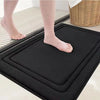 A black plush memory foam bathroom mat is shown with bare feet on a tiled floor.