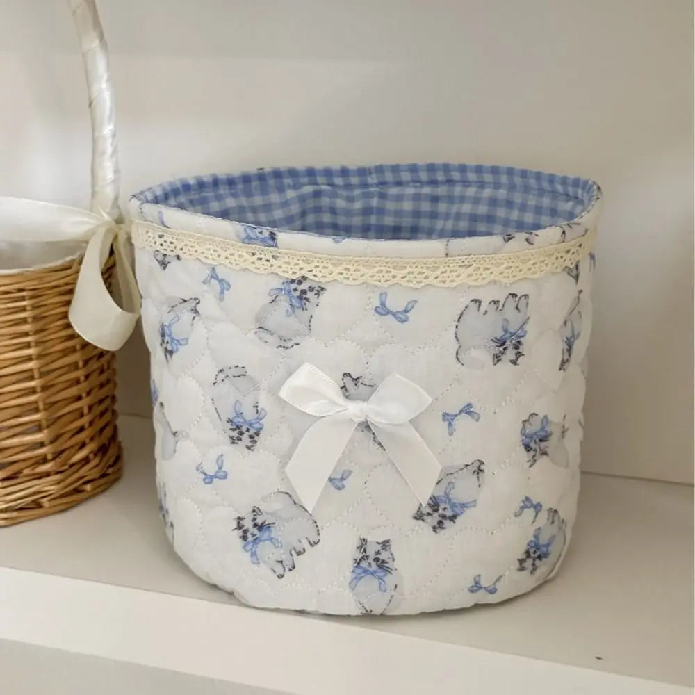 Cesta de almacenamiento acolchada blanca con patrones de mariposa y gato azul, atada con una cinta blanca.