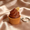 A small, brown, swirl-shaped cake is placed in a paper cup, next to a silver keychain ring.