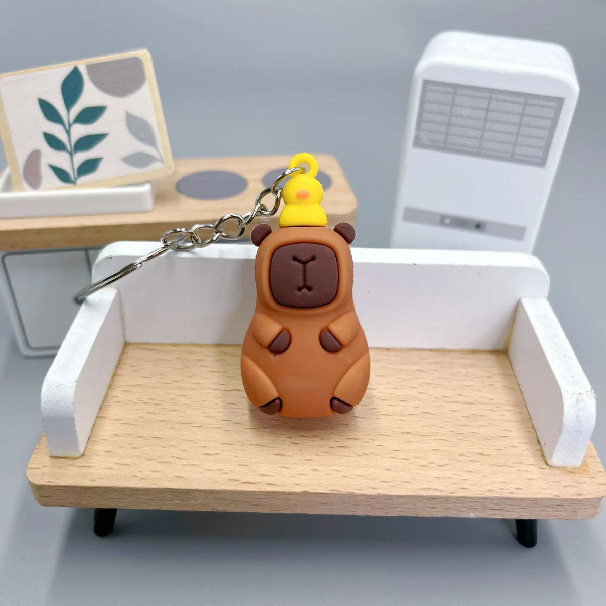 An adorable brown capybara charm with a yellow crown and a silver chain displayed on a wooden shelf.
