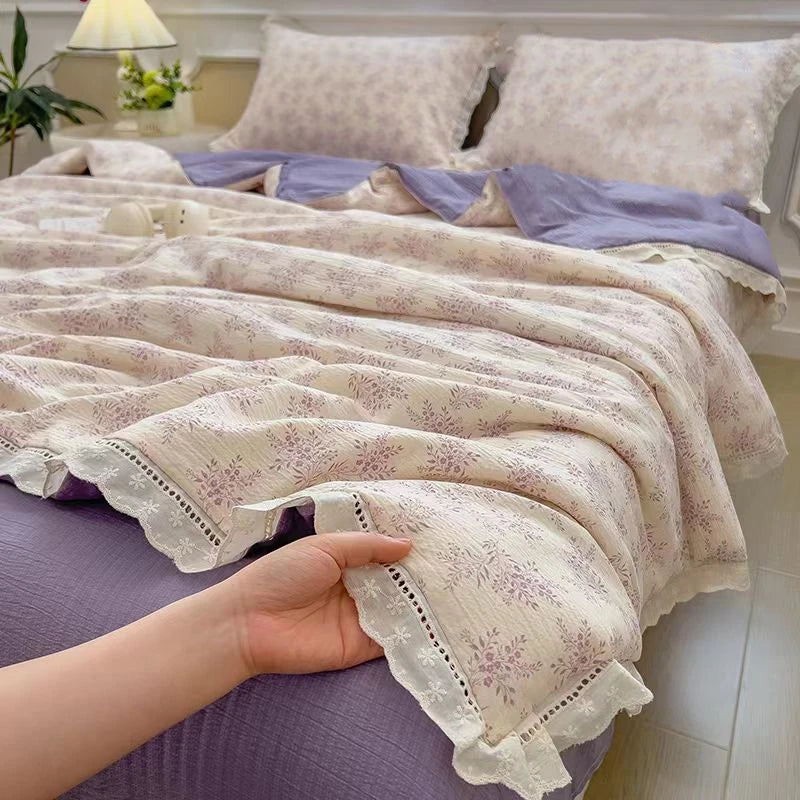 A hand is holding a light purple floral washed cotton gauze summer quilt with white lace trim.