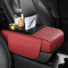 A red quilted car armrest cushion with storage and cup holders placed on a black car seat.