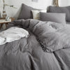A gray duvet cover set with a pom-pom trim is displayed on a bed, featuring matching pillows and a white throw.