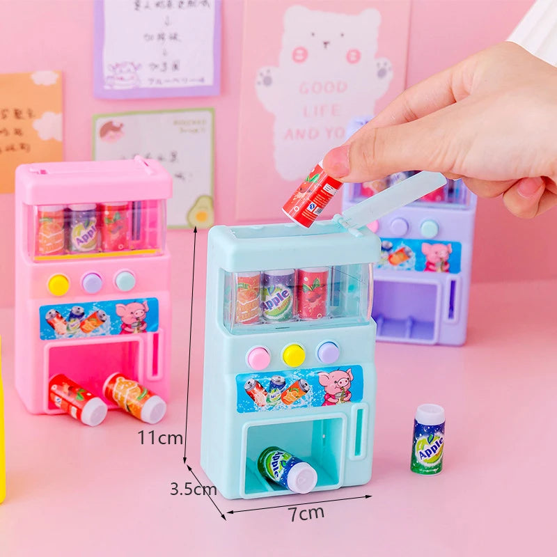 A pink interactive vending machine toy with slots for play coins and drinks is held by a hand against a pink background.