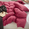 A pink quilted duvet cover set with pom-pom trim resting on a white bed with a gray throw blanket.