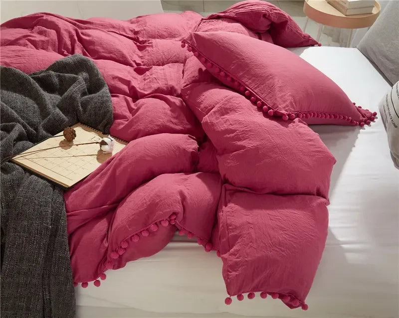 A pink quilted duvet cover set with pom-pom trim resting on a white bed with a gray throw blanket.