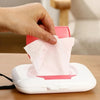 A hand is dispensing pink facial wipes from a white dispenser box onto a wooden surface.