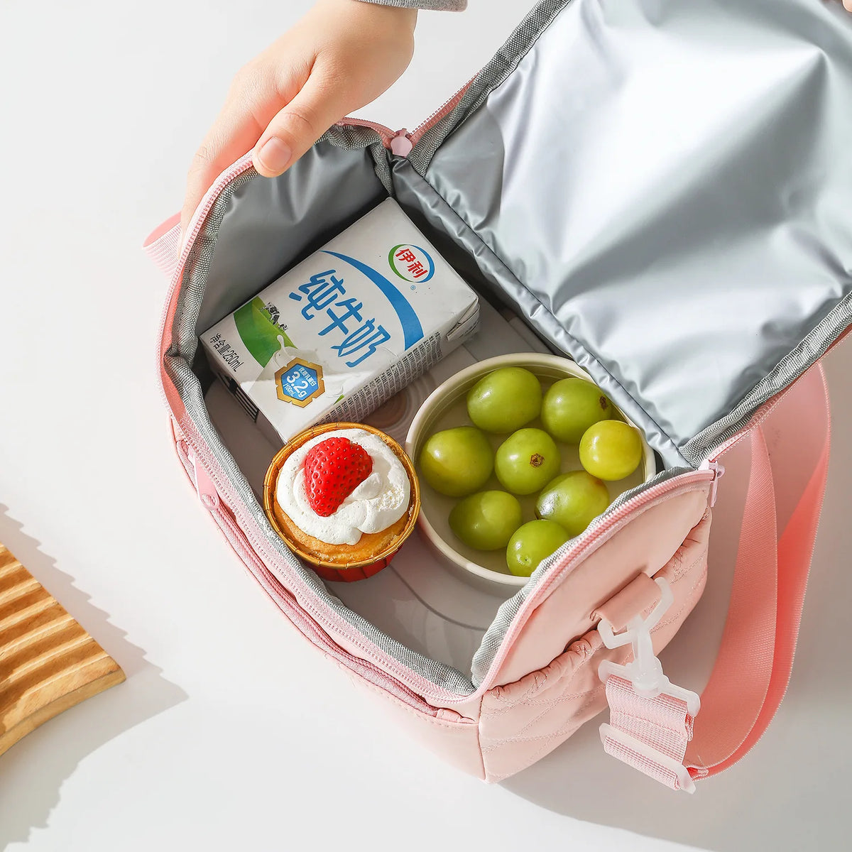 A hand is opening a pink insulated lunch bag to reveal a packaged strawberry dessert and green fruit.