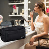 A woman is holding a black travel toiletry bag while sitting at a counter in a well-lit room.