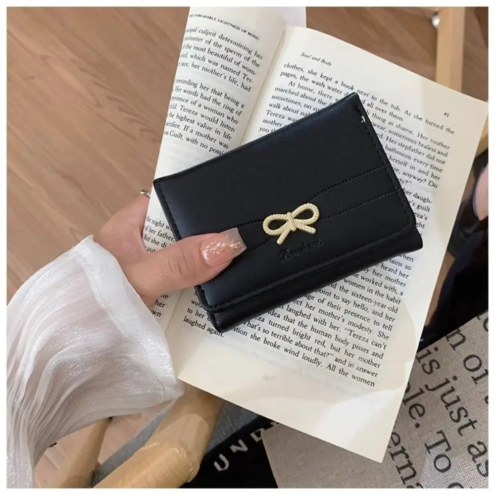 A black tri-fold wallet with a gold bow detail is held by a hand against a background of printed paper.