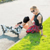 A blonde woman wearing sunglasses is sitting on a grassy slope, showing off a black knee, elbow, and wrist protection set.