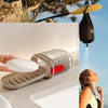 Eine Hand hält eine silberne, dichtende Reise-Seifenschale neben einer weißen Seifenplatte auf einer hellen Oberfläche.