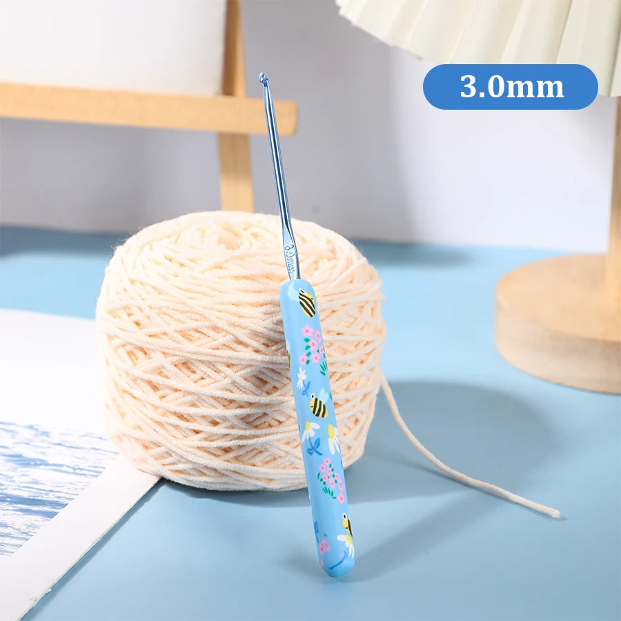 A blue ergonomic crochet hook with a bee design is shown next to a ball of light-colored yarn.