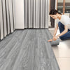 A woman is installing gray wood grain floor tiles in a modern room with sheer curtains.