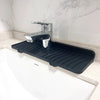 A black silicone faucet splash mat with a chrome faucet is placed on a white countertop next to a sink.