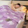 A woman is resting with a purple patterned throw blanket draped over her on a light-colored bed.