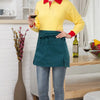 A woman wearing a yellow shirt and dark green half apron with pockets standing in a kitchen setting.