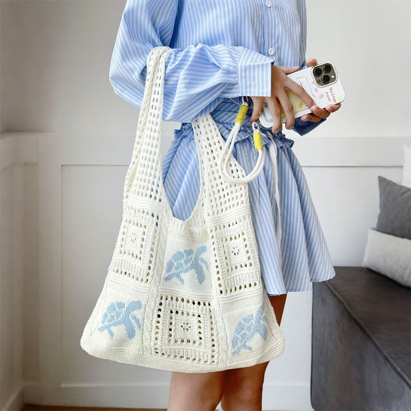 A person holds a white handwoven knit tote bag with blue floral embroidery against a light background.