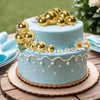 Zwei-stöckiges hellblaues Kuchen mit goldenen Kugendekorationen und weißen Blumenakzenten