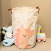 A white fabric storage basket with a pink unicorn design and a yellow-and-pink stuffed animal next to it.