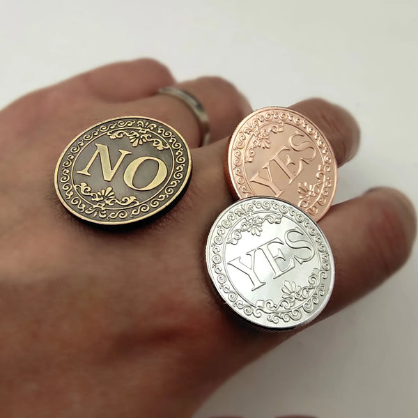 A hand holds three double-sided metal decision coins engraved with NO, YES, and YES on gold, rose gold, and silver finishes.