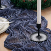 A dark blue crinkle gauze cheesecloth table runner draped on a wooden surface with a lit white candle and greenery.