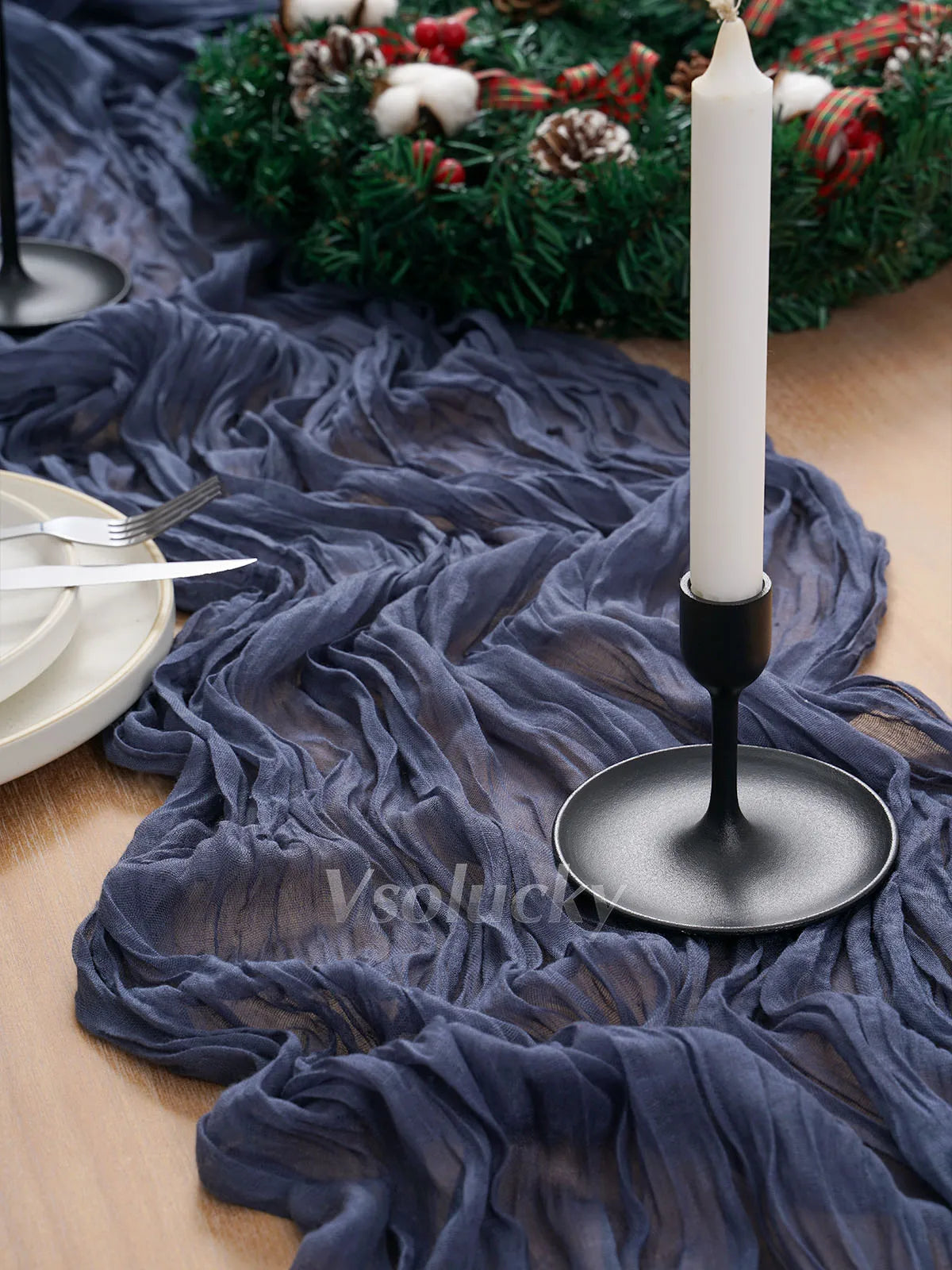 A dark blue crinkle gauze cheesecloth table runner draped on a wooden surface with a lit white candle and greenery.