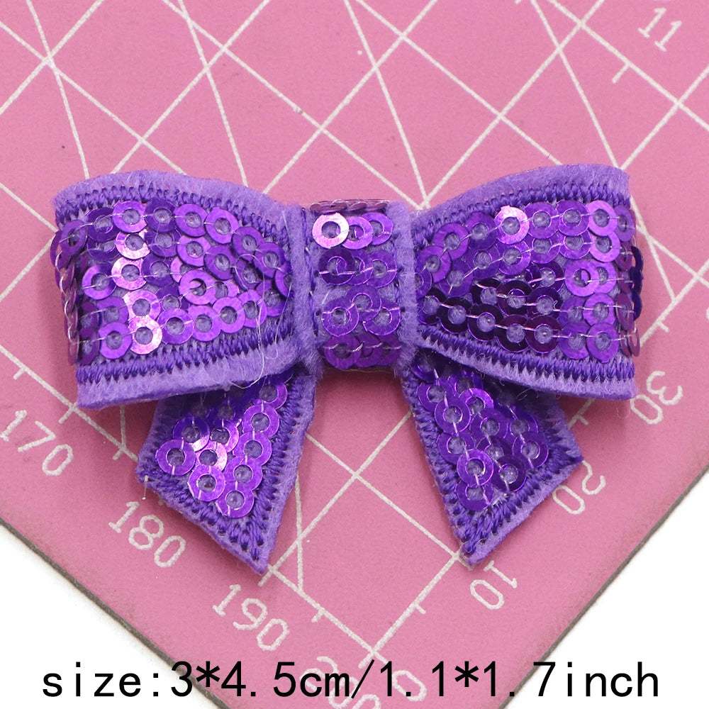 Purple sequin bow patches for DIY crafts shown on a pink grid background