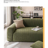 A green quilted sofa cover with a beige throw pillow and a laptop on a dark wooden table in a bright living room setting.