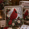 A framed piece of Christmas craft art featuring a red bird made from fabric and greenery on a snowy background.
