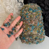 A hand is holding a skein of hand-dyed variegated cotton blend yarn with green and blue fibers.