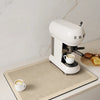 A white Smeez coffee maker sits on a beige counter mat with a white cup of coffee.