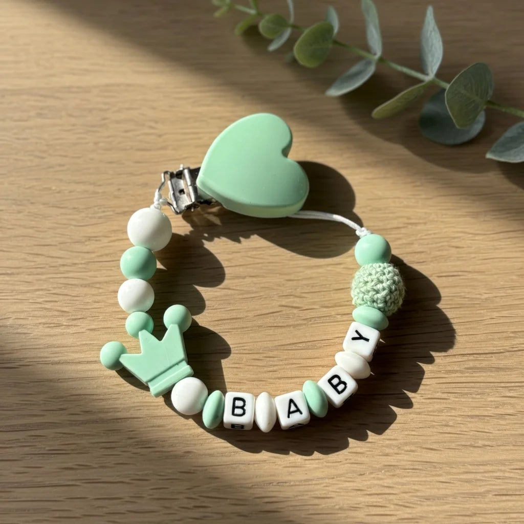 A light green heart-shaped pacifier clip with a string of white and green beads and a crown charm resting on a wooden surface