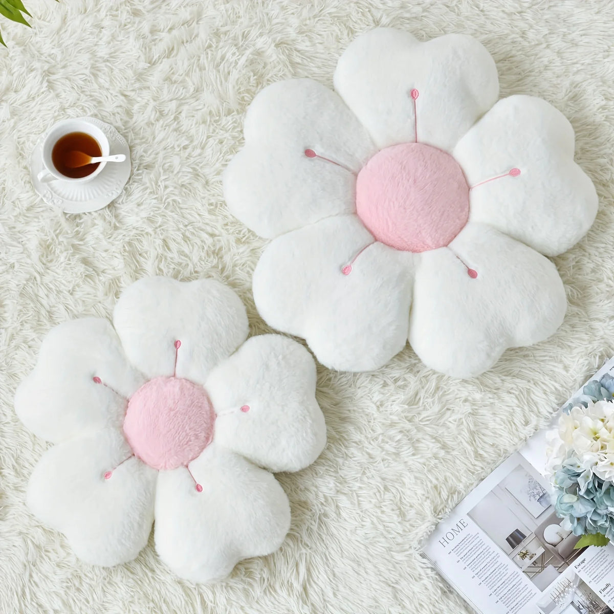 A white plush daisy flower throw pillow with pink accents resting on a cream-colored fluffy surface.