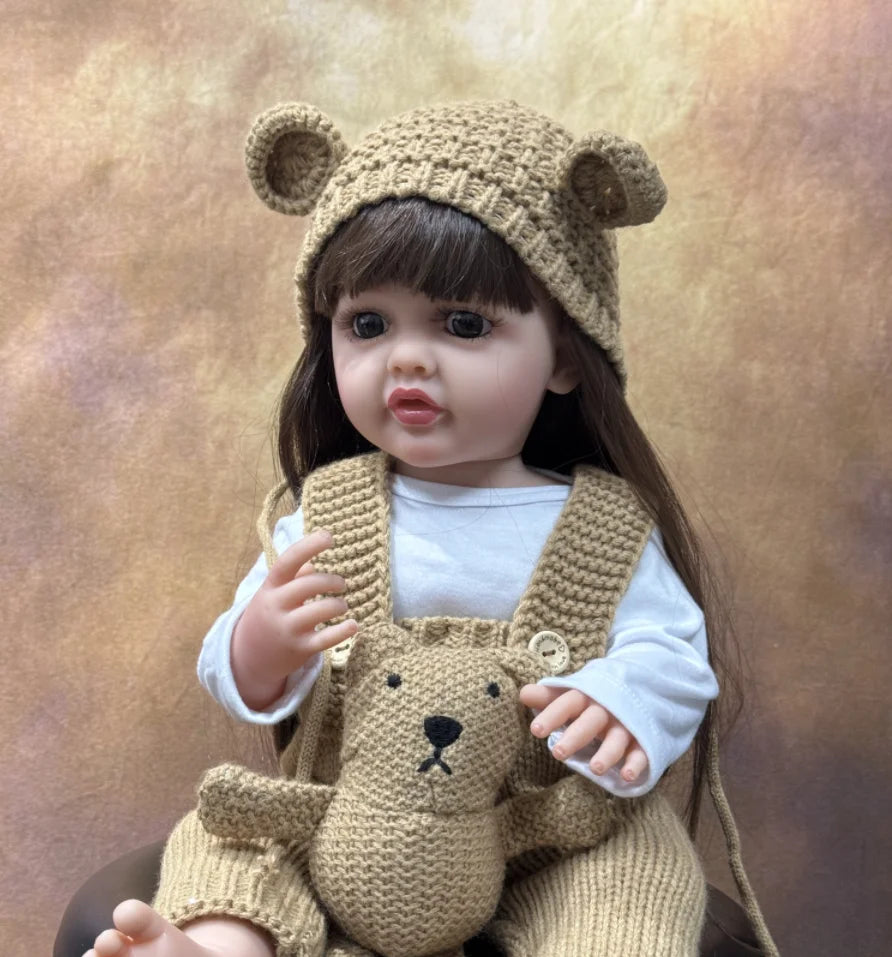 A baby doll wearing a knitted bear hat and overalls, with long dark hair.