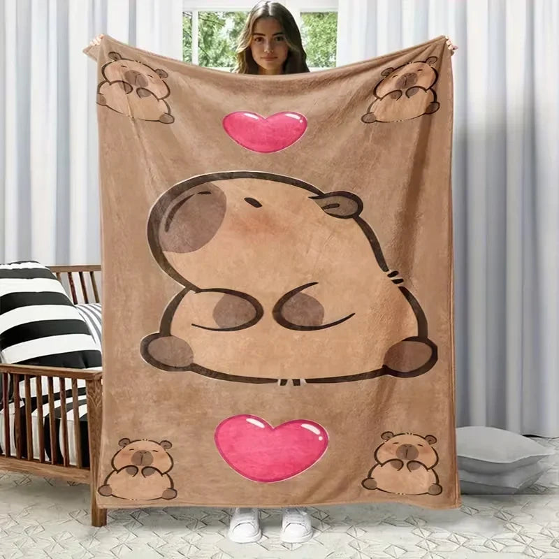A tan flannel throw blanket featuring a large cartoon capybara face and pink hearts, displayed against a white background.