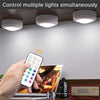 A hand holds a white remote control device used to simultaneously control multiple lights mounted on a ceiling.