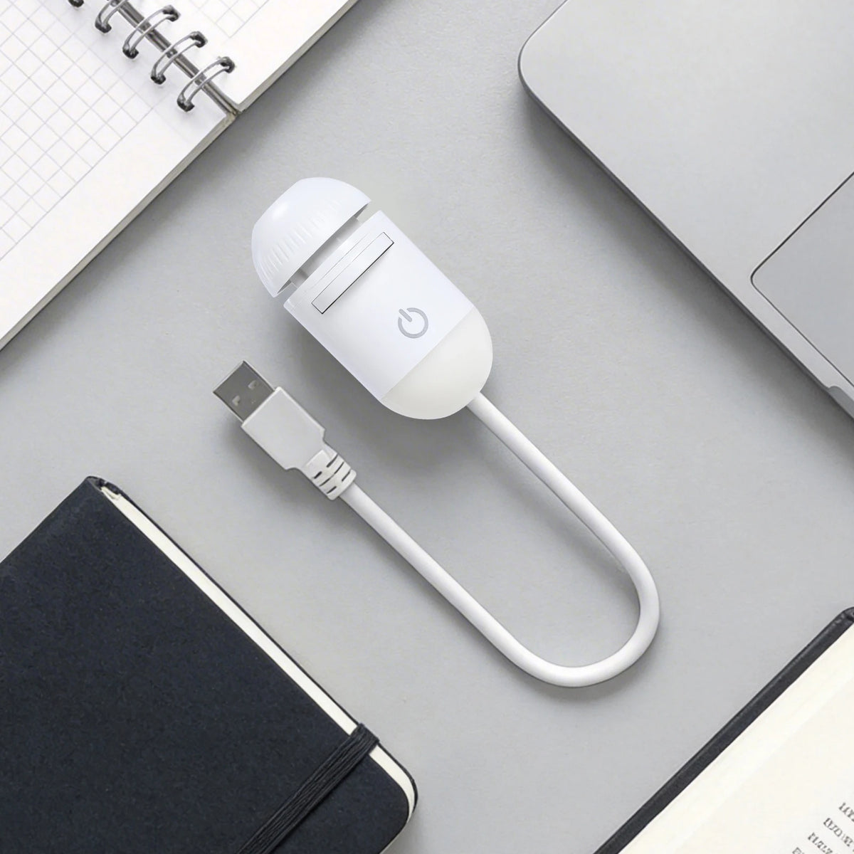 A white 2-in-1 mini projector night light with a flexible cord is shown on a gray surface next to a notebook and a laptop.