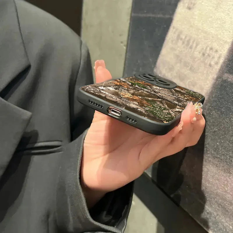 A hand holds a black silicone phone case with a forest camouflage pattern against a dark background.