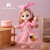 A pink fashion doll with long hair wearing a pink checkered outfit standing next to wooden furniture.