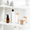 A white bathroom organizer set featuring a glass bottle, a white bottle, and a wooden soap dispenser on a shelf.