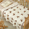A cream cotton gauze baby swaddle blanket with a brown bear print, draped over a wooden crib.
