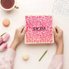 Hands hold a pink stencil card with the word MOM on it, set against a light background.